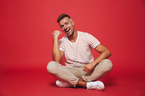 Image of unshaved man 30s in striped t-shirt smiling while sitting on floor with legs crossed isolated over red background - Photo, Image