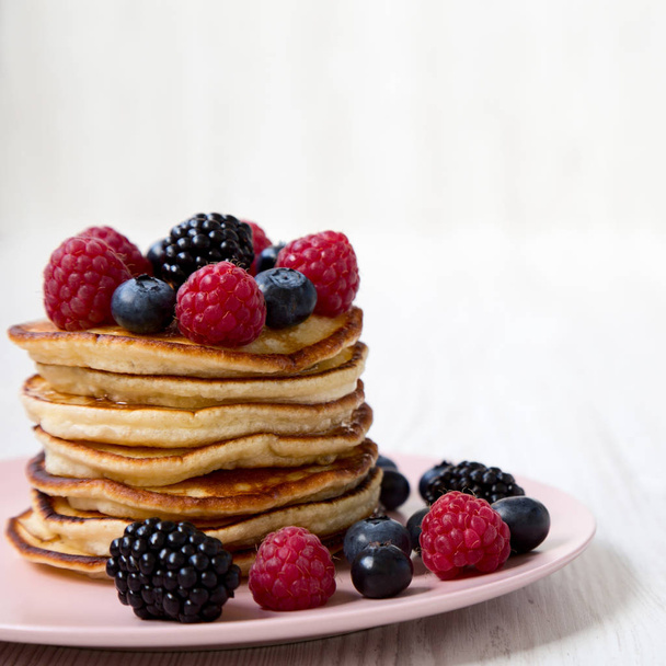 Pancakes with berries on a pink plate over white wooden surface, close-up. Side view. - Photo, image