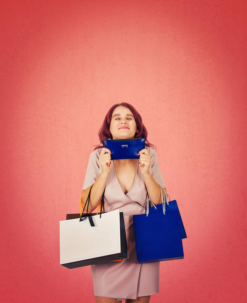 Cheerful girls closed eyes holding a wallet purse the wanted present for holidays among all shopping bags. Shopaholic on sale season isolated over pink background with copy space over head. - Foto, imagen