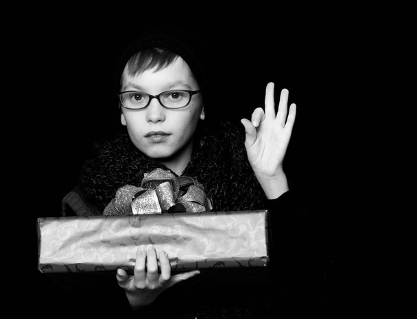 small boy or cute nerd kid in glasses, hat and fashionable knitted scarf on black background holds red present box with golden bow and ok gestur - Foto, immagini