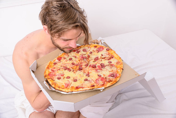 Man likes pizza for breakfast. Bachelors nutrition. Man bearded handsome guy eating cheesy food for breakfast in bed. Guy holds pizza box sit bed in bedroom or hotel room. Food delivery service - Zdjęcie, obraz