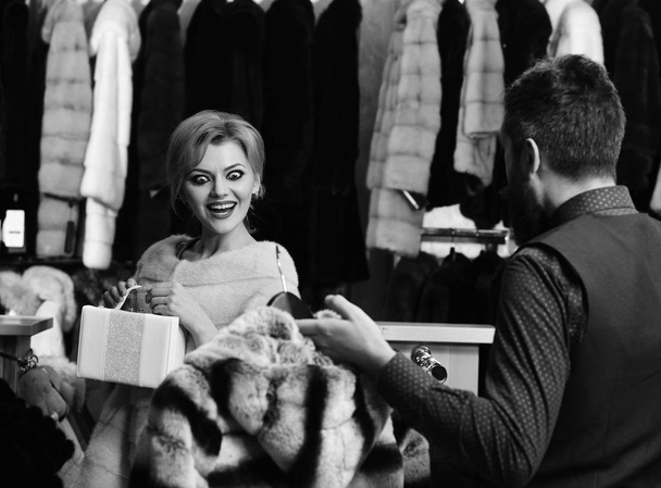 Shop assistant with beard and mustache shows fur coat to lady. Girl in pink fur coat looks at striped fur in male hands. Woman with shocked smile in fashion store. Winter clothing and glamour concept - Foto, Imagem