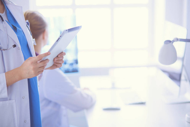 Detailní záběr lékařů ruce drží tablet a kolega sedí u stolu na pozadí. - Fotografie, Obrázek