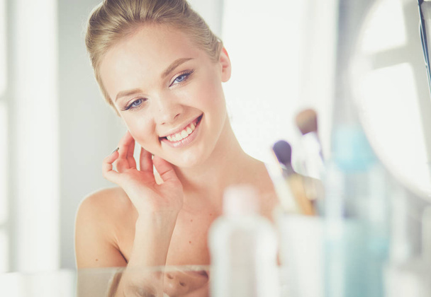 Beautiful young woman is touching her face and smiling while looking at the mirror - Foto, imagen
