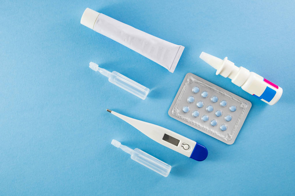 Blister pack of blue pills, ampoules, nasal spary, white ointment tube and digital thermometer on blue background- healthcare and medical concept - Photo, Image