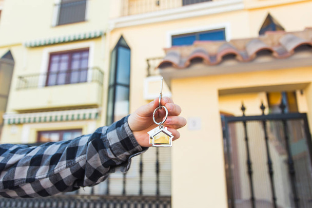 Χέρια που κρατούν τα κλειδιά του σπιτιού στο σπίτι σε σχήμα μπρελόκ μπροστά σε ένα νέο σπίτι. - Φωτογραφία, εικόνα
