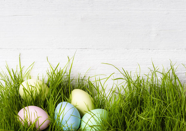 Easter eggs laying in green grass near the old white wooden wall, copy space. Easter holiday decorations - Fotografie, Obrázek