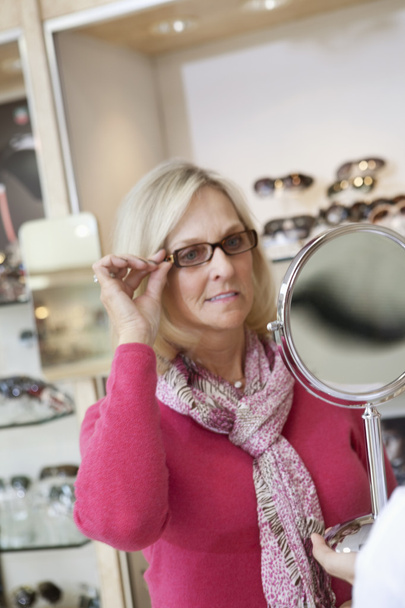 Senior woman trying glasses while looking into hand mirror in store - Fotografie, Obrázek