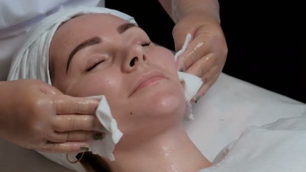Face of a beautiful girl on the procedure of moisturizing and cleaning the skin in the beauty salon. A cosmetologist rubs a woman s neck, chin, forehead, nose and cheeks with a white napkin.    - Footage, Video