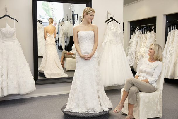 Happy mother looking at young daughter dressed in wedding gown in bridal boutique - Фото, зображення