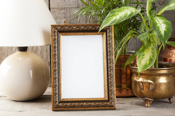 Empty brass picture frame, green plants and table lamp on old wooden gray textured background. Home decor and copy space for text.   - Photo, Image
