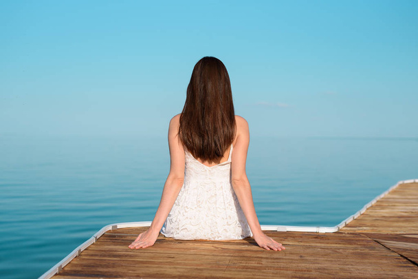 Uma menina de vestido branco sentado no cais e olhando para a distância
 - Foto, Imagem