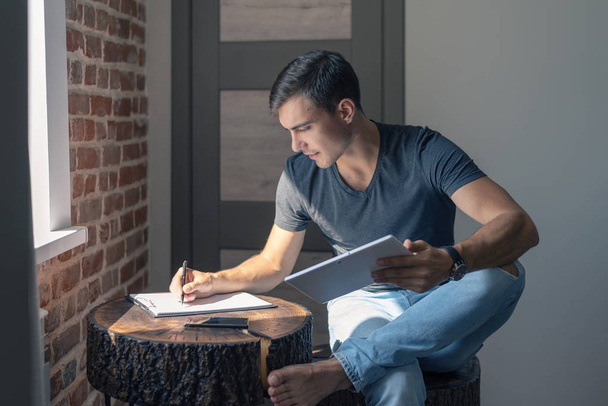 Χαριτωμένο νεαρό άνδρα σε t-shirt και τζιν στο ξύλινο τραπέζι με tablet, γράφει στο τετράδιο, επιχειρηματικό σχέδιο, ελεύθερος επαγγελματίας, εκκίνησης - Φωτογραφία, εικόνα
