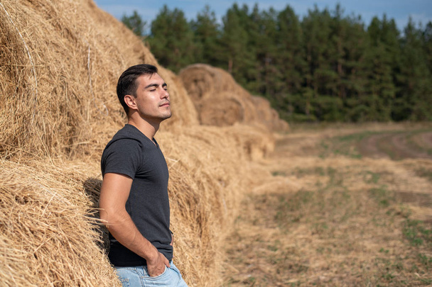 young man in gray t-shirt and jeans stands at haystack, breathing clean air, freshness, rest after work, harvesting, concept, copy space - Foto, afbeelding