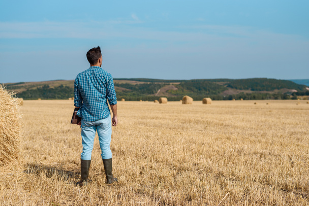 Man farmer agronomist in jeans and shirt stands back in field after haymaking, with tablet looking into the distance. Rural business, agricultural industry, freedom after work, concept - Photo, Image