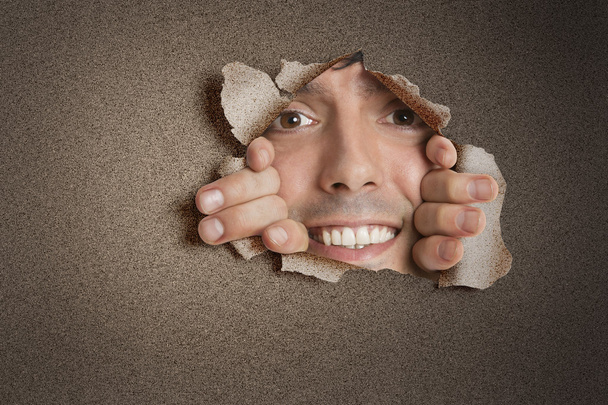 Portrait of a happy young Hispanic man peeking from ripped paper hole - Zdjęcie, obraz