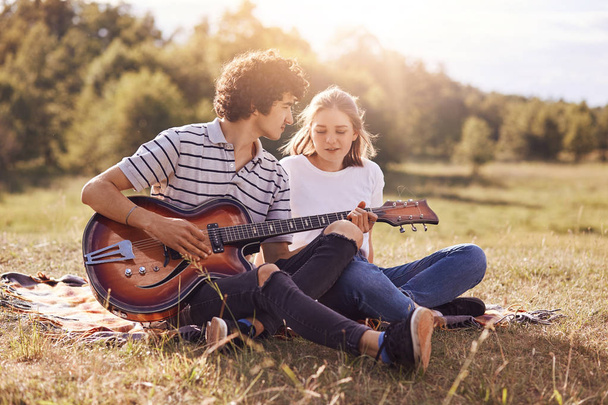 Lovely couple of teenagers spend free time together, have date, enjoys peaceful atmosphere outdoor in meadow. Romantic guy plays guitar and sing songs to his girlfriend, expresses great love - Фото, зображення
