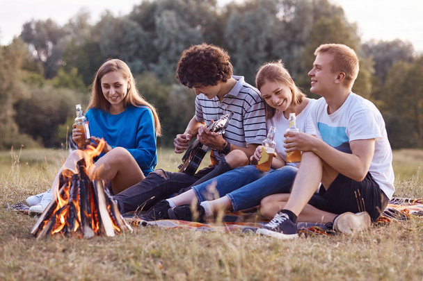 Joyful teenagers sit closely to each other on ground near fire, have picnic together, play acoustic guitar, celebrate something, have outdoor party, drink cold energetic drinks. Leisure concept - Foto, Imagem