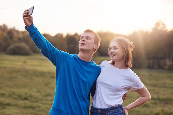 Photo of joyful lovely couple stand closely to each other, look positively into screen of smart phone, make selfie, have good recreation time on nature, enjoy togetherness. Vacation and rest concept - Фото, изображение