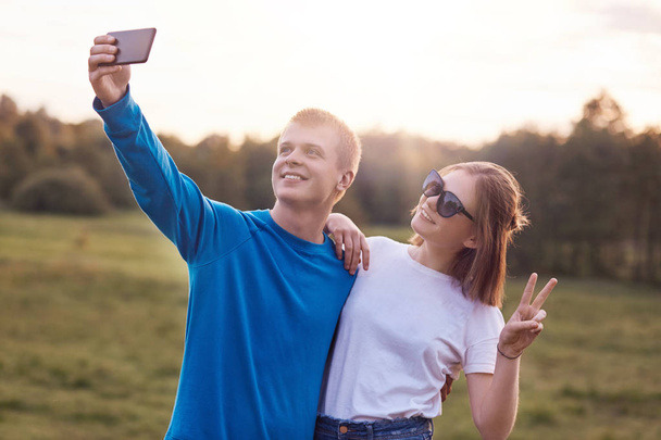 Outdoor shot of joyful teenagers embrace and stand next to each other against nature background, make selfie with modern smart phone for social networks, show peace sign, have happy expressions. - Φωτογραφία, εικόνα