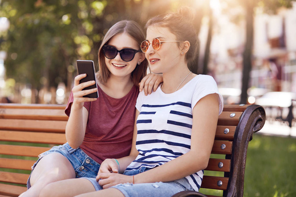 Photo of happy teenage girls message in social networks, use smart phone for entertainment, wears trendy sunglasses, pose on wooden bench outdoor, connected to wireless internet. Leisure concept - Foto, Imagem