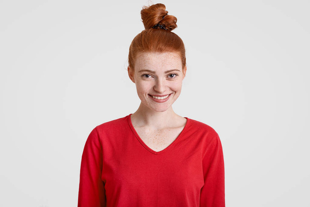 Cheerful attractive teenager with ginger hair knot, freckled skin, dressed in casual red sweater, being in good mood as has date with boyfriend, models against white background. Beauty concept. - Фото, изображение