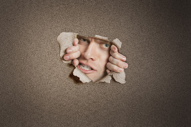 Portrait of a aggressive mid adult man peeking from ripped white paper hole - 写真・画像
