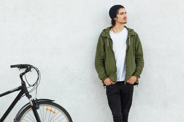 Youth and cycling concept. Stylish guy dressed in hat, green jacket, keeps hands in pocket, stands near sport bicycle, focused aside with contemplative look, thinks abut new travels by bike. - Fotoğraf, Görsel