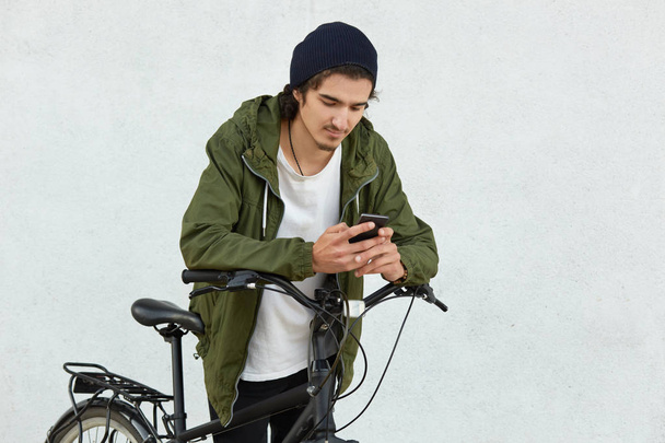Stylish youngster in black hat and jacket with hood, has active lifestyle, covers long distances with his sport bicycle, has break, holds smart phone in hands, reads notification, types feedback - Fotoğraf, Görsel