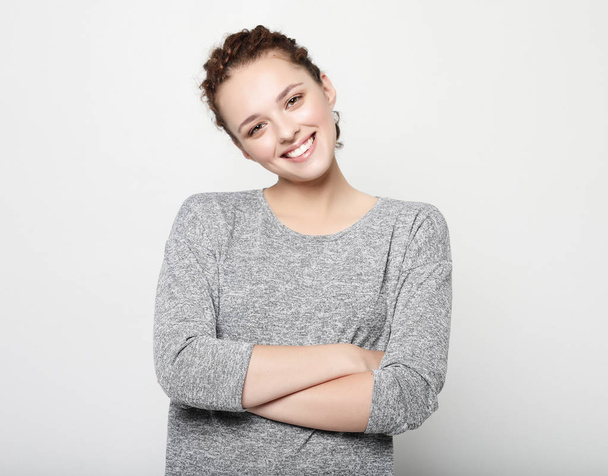 beautiful young woman with curly hair smiling gently while listening to interesting conversation, wearing grey long-sleeved sweater, keeping arms folded. - Foto, Imagen