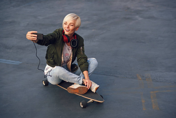 Pretty blond girl with headphones, making self photo on smartphone while sitting on the longboard at sunset. - Φωτογραφία, εικόνα