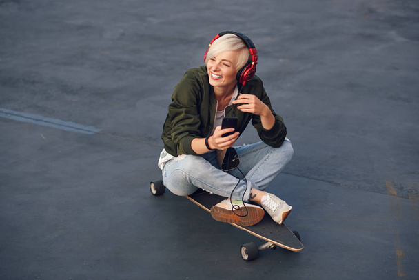 Young smiling blond girl listening music in headphones and using smartphone while sitting on the longboard at sunset. - Fotoğraf, Görsel