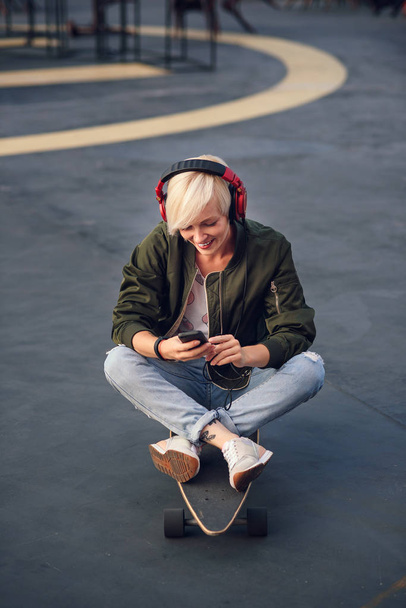 Joven chica rubia sonriente escuchando música en los auriculares y utilizando el teléfono inteligente mientras está sentado en el longboard al atardecer
. - Foto, Imagen