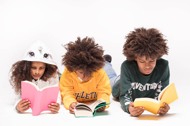 Happy smiling three afro children in colorful clothes laying on the floor, reading books. - 写真・画像