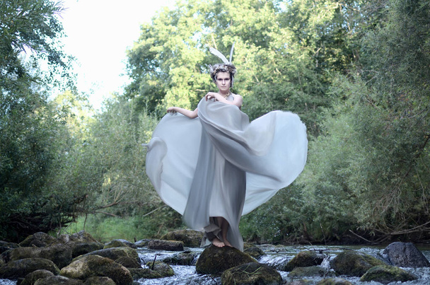 jonge vrouw in een grijze jurk van zijde, een hoofdtooi van veren en een ketting van zee schelpen staat op de rotsen onder de stroom van een onstuimige rivier en golven van de trein van haar jurk - Foto, afbeelding