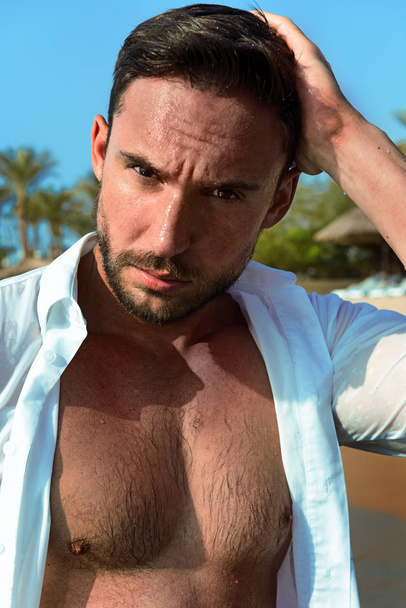 closeup of brutal handsome man in white wet shirt sitting on a sand beach enjoying summer, a muscular male model poses naked for magazine with a smirk in his eyes. Romantic bearded macho - Photo, Image