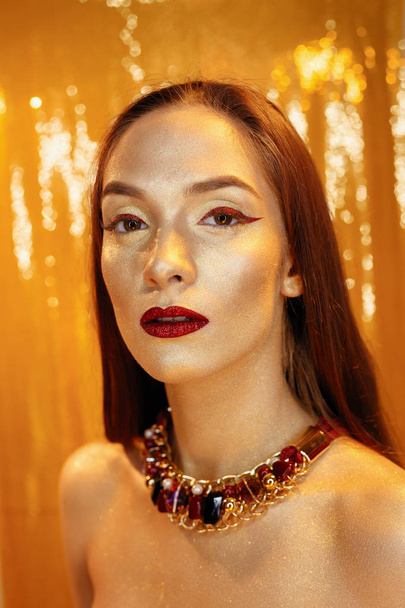 Magic Girl Portrait in Gold. Golden Makeup, close-up portrait in studio shot, color. Beauty Model Girl with perfect bright make-up, red lips, golden maroon jewellery. Sexy lady makeup Holiday party. - Fotó, kép