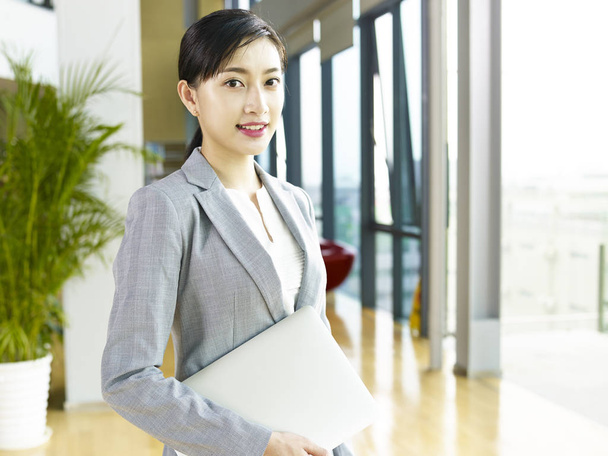 portrét mladé Asijské obchodní ženy stojící v hale moderní kancelářská budova držení notebooku při pohledu na fotoaparát s úsměvem. - Fotografie, Obrázek