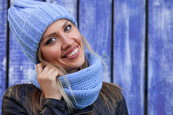Portrét krásné mladé modelu v modré pletené čepice, stojící poblíž modré zdi na podzimní den. Teplé podzimní foto. Žena s úsměvem a podívat se na kameru, radostné náladě. - Fotografie, Obrázek