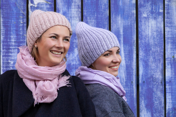 Portrét dvou krásných mladých modelů v modré a růžové pletené čepice, stojící poblíž modré zdi na podzimní den. Teplé podzimní foto. Žena s úsměvem a podívat se na kameru, radostné náladě. - Fotografie, Obrázek