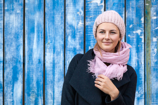 Portrait of a beautiful young model in pink knitted hat standing near blue wall on an autumn day. Autumn warm photo.Woman smiling and look at the camera, joyful cheerful mood. - Fotografie, Obrázek