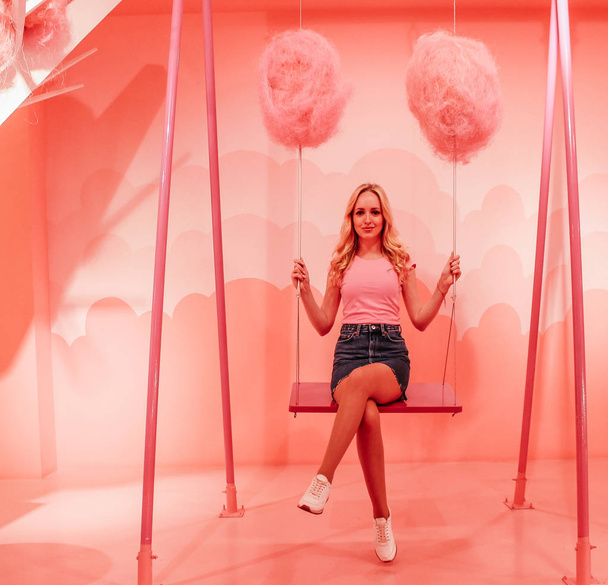 Young cute blonde girl is riding on a swing chain on a bright pink background in the form of clouds. - Foto, Imagem
