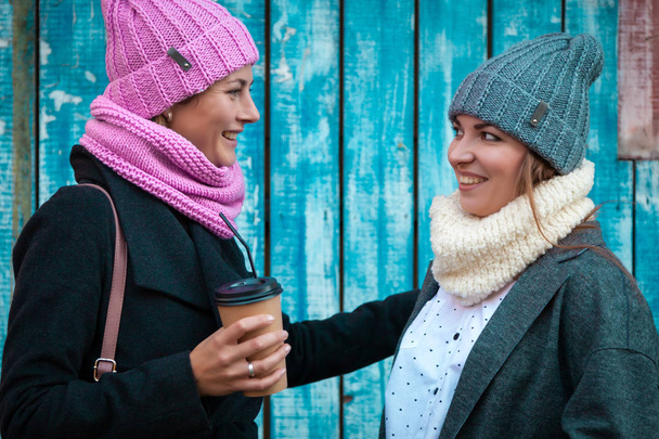 Przyjaciele młodych kobiet w stylowe ubrania przytulić, czat i napić się kawy, aby przejść wokół niebieską ścianą. Pojęcie trybem życia i rekreacji na świeżym powietrzu w jesieni - Zdjęcie, obraz
