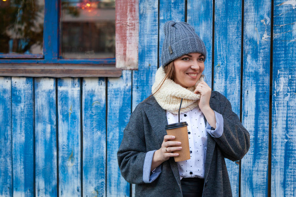 Een jonge vrouw in een grijze gebreide muts, de grijze vacht en de bluesses geniet van stad, poses en drinken koffie te gaan rond blauwe muur. Het concept van wonen en outdoor recreatie in de herfst - Foto, afbeelding