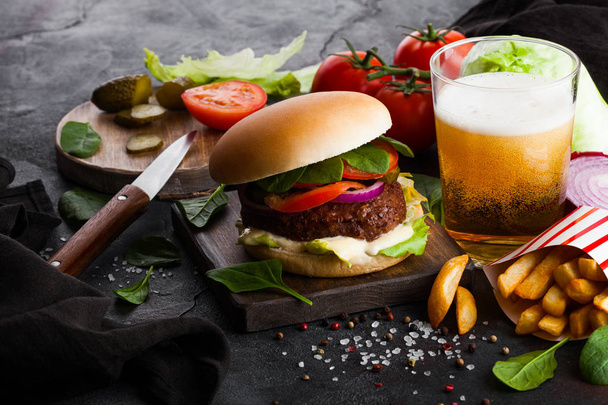 Fresh beef burger with sauce and vegetables and glass of lager craft beer with potato chips fries on stone kitchen background. - Foto, imagen