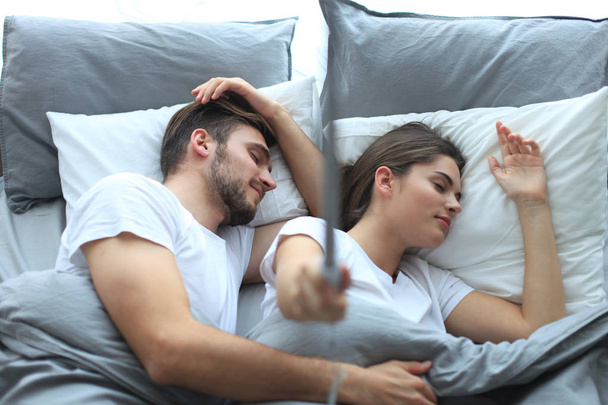 Beautiful young loving couple sleep and making selfie with stick in the bed. - Фото, зображення