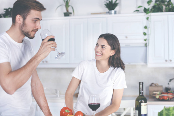 Beautiful young couple is drinking wine and smiling while cooking in kitchen at home. - Φωτογραφία, εικόνα