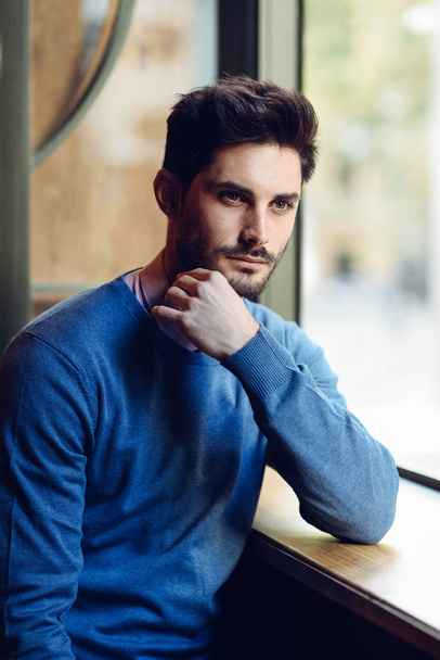 Thoughtful man with blue sweater with lost look near a window in a modern pub. Bearded guy with modern hairstyle. Life style concept. - Photo, Image