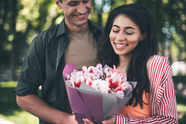 Favorite flowers. Positive emotional young woman smiling happily while standing next to her cheerful boyfriend and looking at the bouquet of flowers - 写真・画像