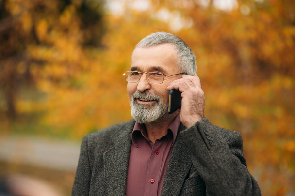 Egy jóképű idős férfi szakáll, szemüveg segítségével a telefon. Őszi séta a parkban - Fotó, kép